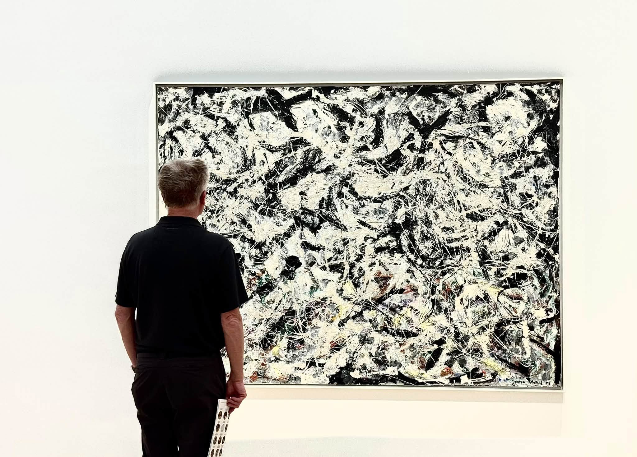 Tom, seen from behind in a black shirt, standing before a large black, white, and gray Jackson Pollock drip painting in a white gallery