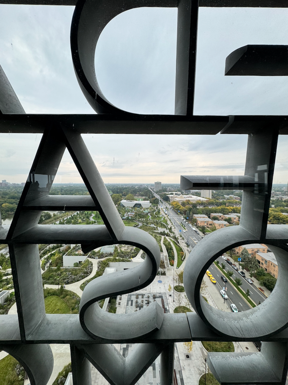 View through large sculpted letters in a window looking down on the green paths of Jackson Park and a tree-lined boulevard below
