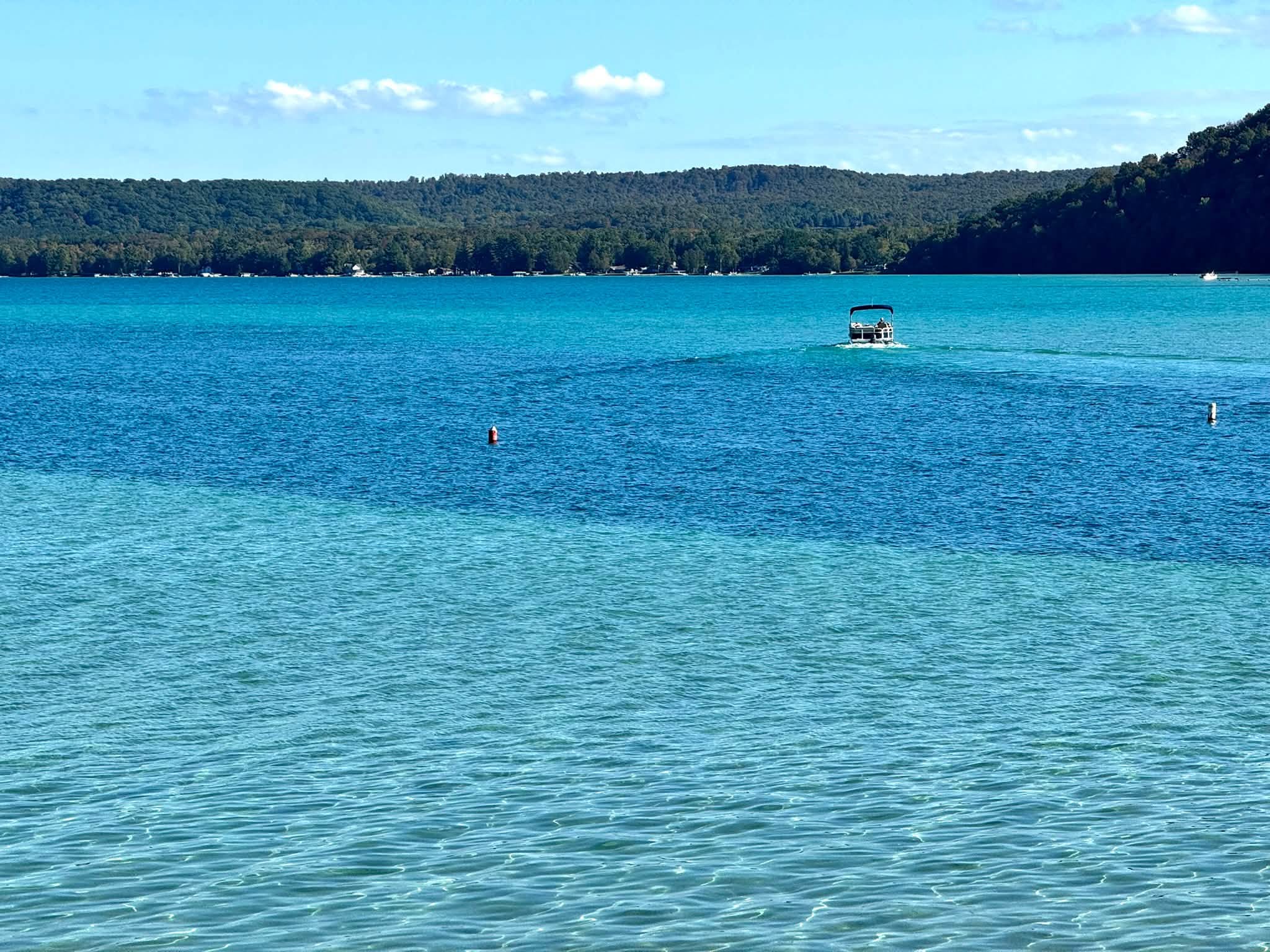 A pontoon boat crossing Glen Lake’s clear turquoise shallows and deep blue water, with forested hills and lakeside cottages beyond