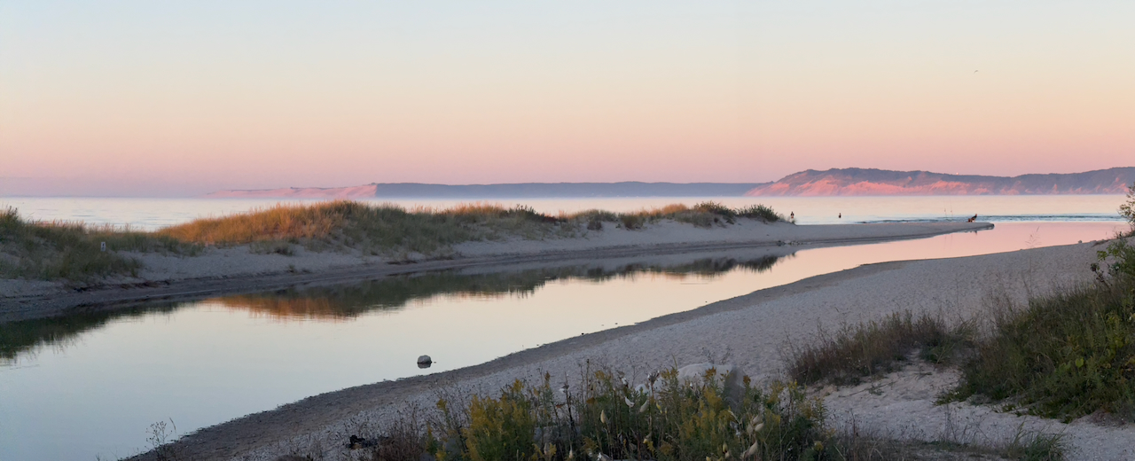 Pink and gold sunset light over the mouth of a river winding past grassy dunes into Lake Michigan, with distant bluffs on the horizon